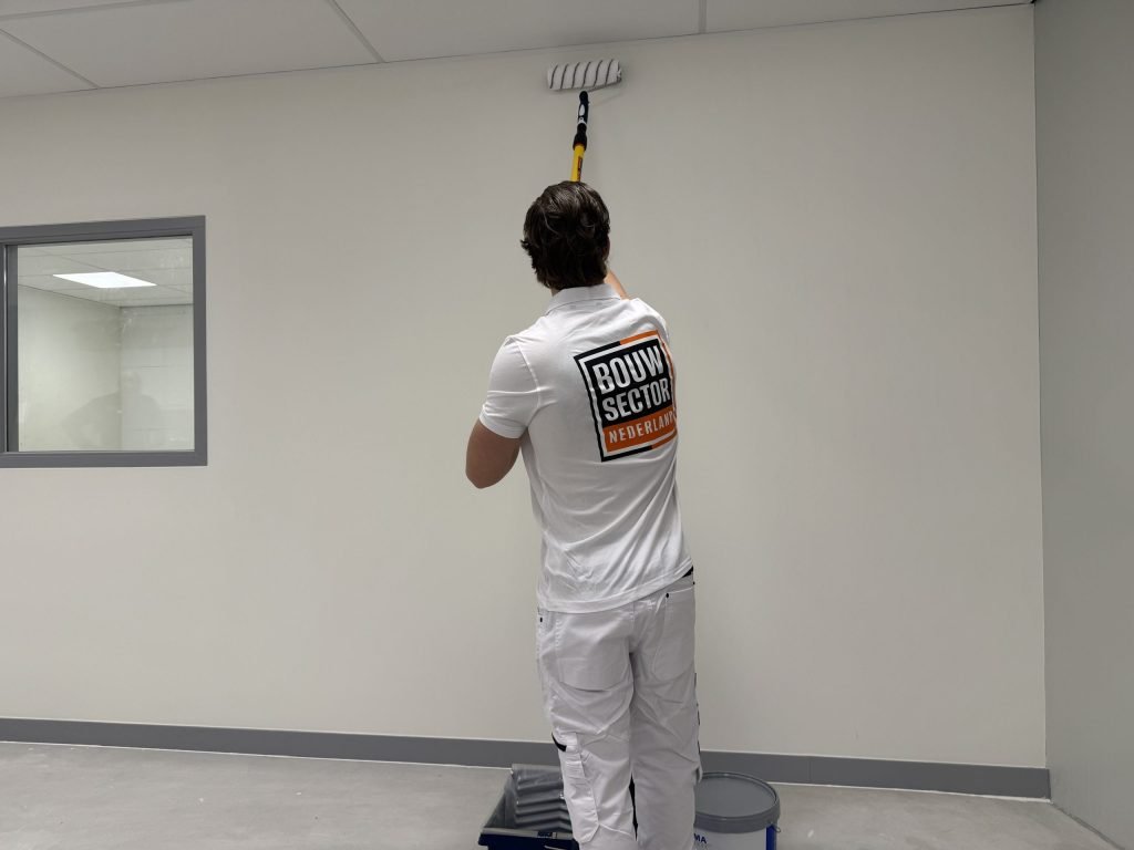 Schilder rolt de bovenrand van een witte wand vlak onder het plafond, met verfbak en emmer aan zijn voeten