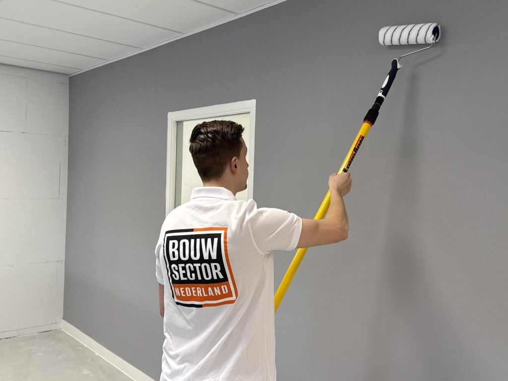 Schilder van Bouwsector Nederland zet met een lange roller grijze verf hoog op een wand naast een deur