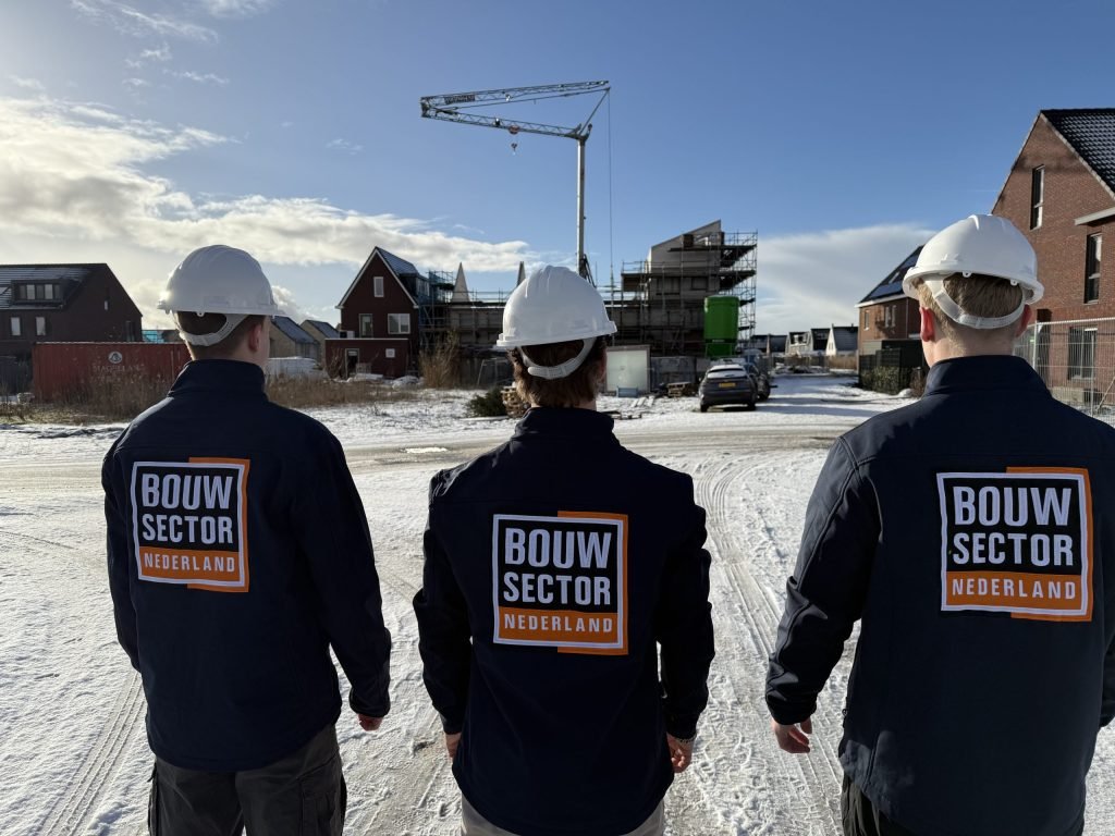 Drie medewerkers van Bouwsector Nederland met witte helm en donkere jas, gezien op de rug, in een besneeuwde straat met bakstenen huizen