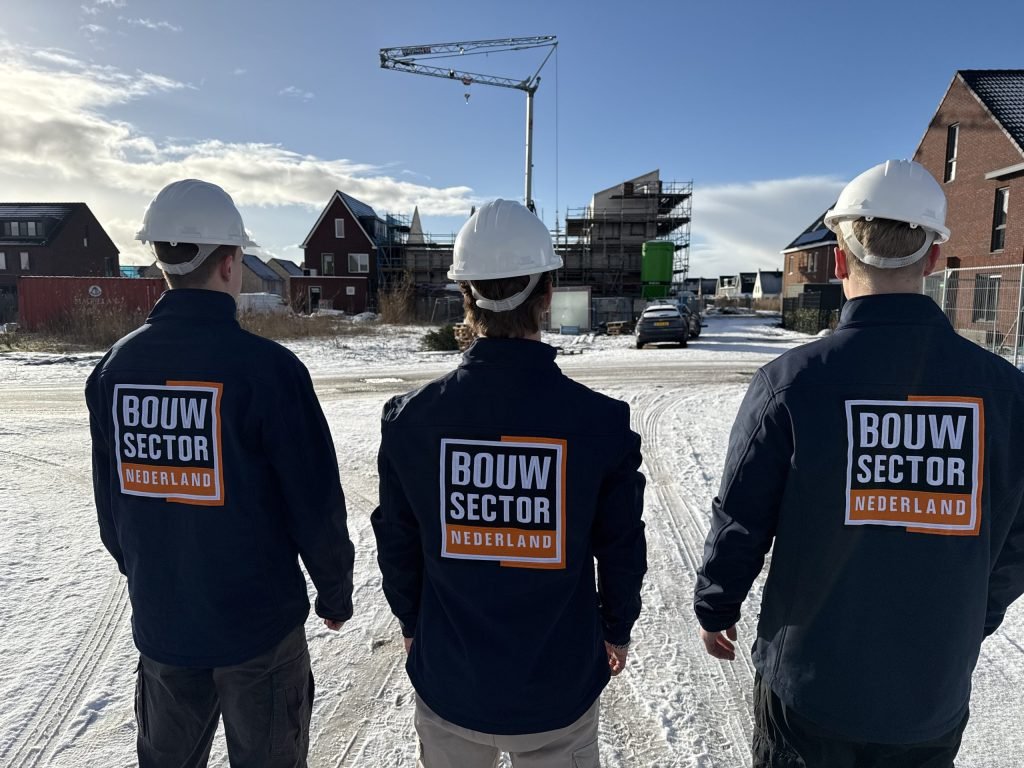 Drie medewerkers van Bouwsector Nederland met witte helm en logo op de jas lopen door een besneeuwde wijk met nieuwbouw en een bouwkraan